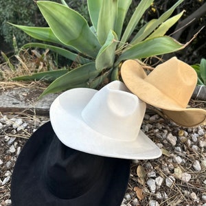 May include: Three felt hats in black, off-white, and tan colours. Each hat features a pinched crown and a wide, curved brim. The hats are stacked on top of each other, showcasing their smooth texture and structured shape in a neutral colour palette.
