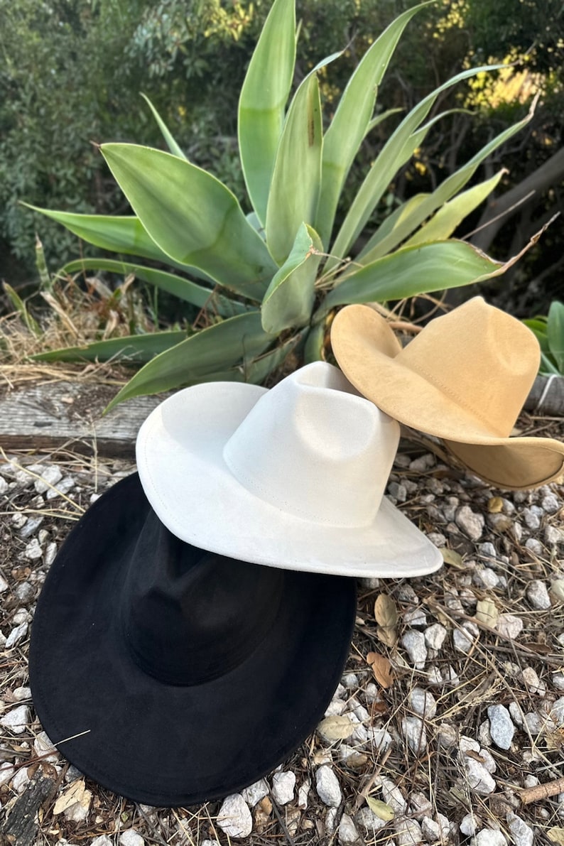 May include: Three felt hats in black, off-white, and tan colours. Each hat features a pinched crown and a wide, curved brim. The hats are stacked on top of each other, showcasing their smooth texture and structured shape in a neutral colour palette.