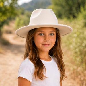 May include: A cream-coloured fedora hat with a structured crown and a wide, flat brim, paired with a simple white ribbed short-sleeve shirt. The hat features a matching tonal band around the base of the crown, creating a clean and uniform appearance.