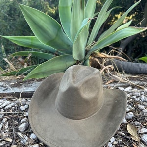 May include: A solid olive-green cowboy hat with a pinched crown and a wide, curved brim. The hat features a smooth, matte-textured finish and a stitched edge detail along the perimeter of the brim, presenting a structured and uniform appearance.
