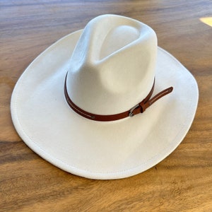 May include: Cream-colored cowboy hat with a brown leather band and buckle. The hat has a wide brim and a rounded crown. The leather band is stitched with white thread. The hat is sitting on a wooden surface.