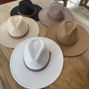 May include: Five felt fedora hats with leather bands in various colors, including black, beige, brown, and white, displayed on a wooden surface.