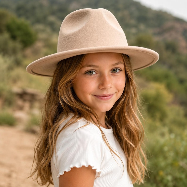 May include: A beige felt fedora hat with a structured crown and a wide, flat brim. The hat features a smooth, uniform texture and a neutral light tan colour, designed with a classic pinched crown shape and a clean, minimalist aesthetic throughout.