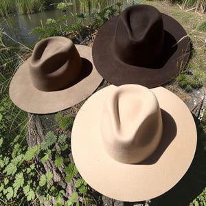 May include: Three felt hats in varying shades of brown and beige are displayed outdoors. The hats have a classic fedora style with a wide brim and a pinched crown. The hats are arranged on a weathered wooden surface, with greenery in the background.