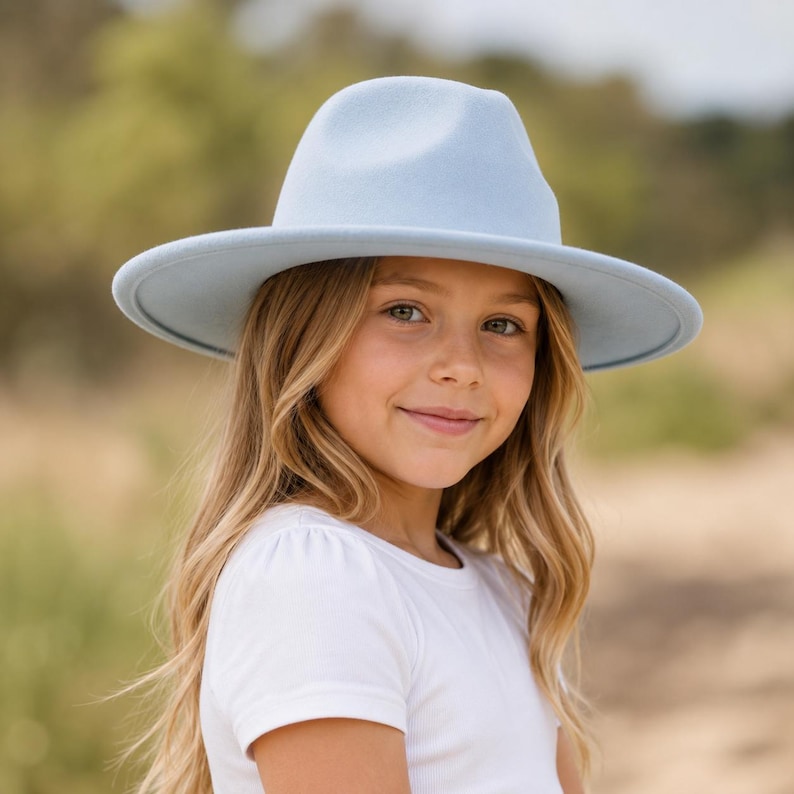 May include: A light blue fedora hat with a structured crown and a wide, flat brim. The hat features a smooth, felt-like texture and a matching tonal band around the base of the crown, providing a clean and uniform appearance in a soft pastel shade.