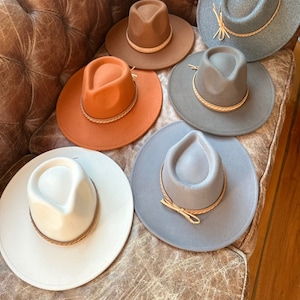May include: Six felt fedora hats with leather bands in various colors, including gray, brown, white, and orange, arranged on a brown leather couch.