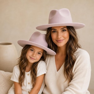 May include: Two matching light lavender fedora hats with a structured crown and a wide, flat brim. The hats feature a smooth, felt-like texture and a clean, minimalist design. These hats are worn by two individuals, one adult and one child.