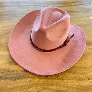 May include: A dusty rose-colored cowboy hat with a wide brim and a brown leather band. The hat has a suede-like texture and a classic western design. The leather band is secured with a buckle and white stitching.