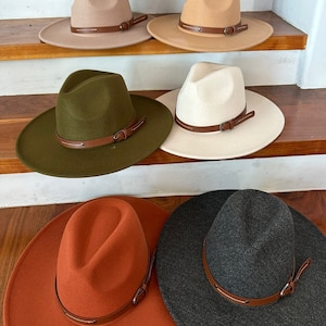 May include: Seven felt fedora hats with leather bands in various colors, including brown, beige, green, white, red, orange, and gray. The hats are arranged on a set of wooden stairs.