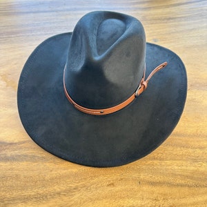 May include: A black fedora-style hat with a wide brim and a brown leather band. The hat has a suede-like texture and a classic, stylish design. The leather band is secured with small metal rivets.