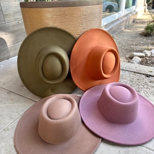 May include: Four felt hats in different colors: olive green, burnt orange, light brown, and dusty rose. The hats have a wide brim and a tall, rounded crown.
