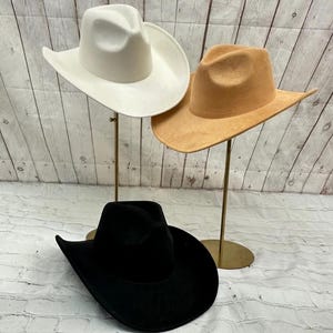 May include: Three cowboy hats displayed together. One hat is solid white, one is solid tan, and one is solid black. Each hat features a high crown with a central crease and a wide, curved brim. The hats are positioned on two metal stands.