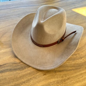 May include: A tan cowboy hat with a brown leather band and buckle. The hat has a wide brim and a rounded crown. The hat is made of a soft material and is likely suede or felt.