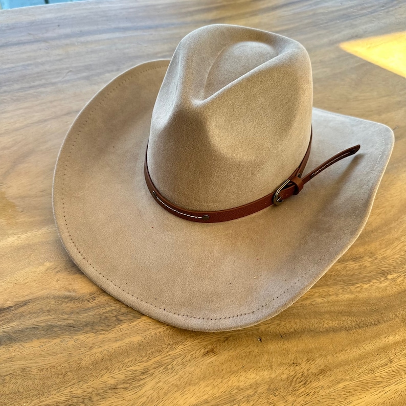 May include: A tan cowboy hat with a brown leather band and buckle. The hat has a wide brim and a rounded crown. The hat is made of a soft material and is likely suede or felt.