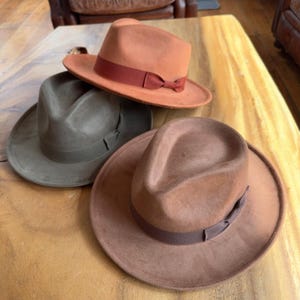 May include: Three felt fedora hats in different colors. The hats are brown, green, and orange. The hats have a ribbon around the crown.