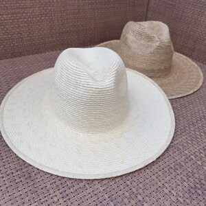 May include: Two straw hats, one white and one tan, are sitting on a woven surface. The hats are both wide-brimmed and have a woven texture.