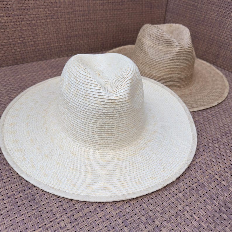 May include: Two straw hats, one white and one tan, are sitting on a woven surface. The hats are both wide-brimmed and have a woven texture.