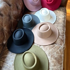 May include: Seven felt hats in various colors, including pink, red, white, gray, beige, black, and green, are arranged on a brown leather couch.