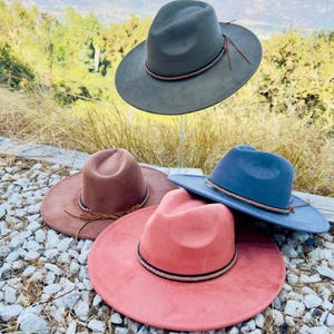 May include: Three felt fedora hats with braided leather bands. The hats are in shades of gray, brown, and pink.