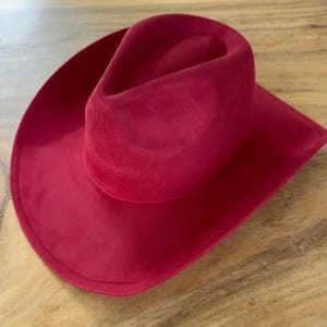 May include: A vibrant red cowboy hat with a smooth, matte texture. The hat features a classic pinched crown design and a wide, curved brim. The material has a soft, suede-like appearance, and the entire piece is a solid, uniform shade of deep red.