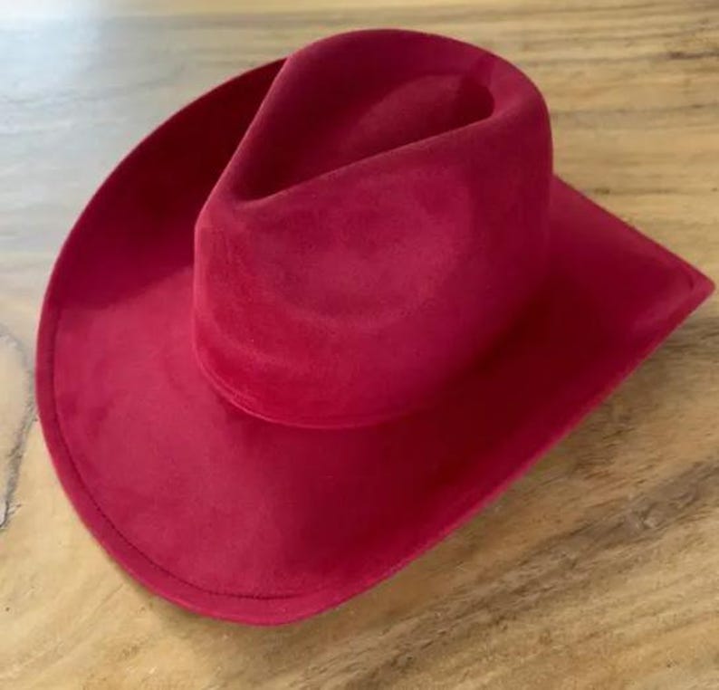 May include: A vibrant red cowboy hat with a smooth, matte texture. The hat features a classic pinched crown design and a wide, curved brim. The material has a soft, suede-like appearance, and the entire piece is a solid, uniform shade of deep red.