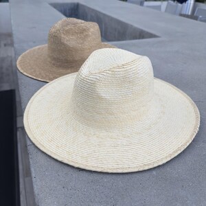 May include: Two straw hats, one beige and one white, are sitting on a gray concrete surface. The white hat has a wide brim and a slightly indented crown.