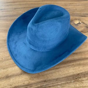 May include: A solid blue cowboy hat with a soft, matte, suede-like texture. The hat features a pinched crown and a wide, curved brim. The design is uniform in colour, showcasing a deep blue hue across the entire exterior surface of the hat.