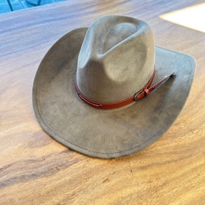 May include: A taupe-colored cowboy hat with a brown leather band and buckle. The hat has a wide brim and a rounded crown. The leather band is secured with white stitching. The hat is made of a soft, suede-like material.