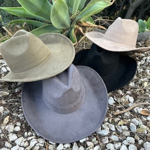 May include: A collection of four wide-brimmed fedora hats in olive green, light beige, black, and dark grey. Each hat features a structured crown with a central crease and a wide, flat brim. The hats are arranged in a cluster, showcasing their smooth, matte-textured finish and consistent design across all four colour variations.