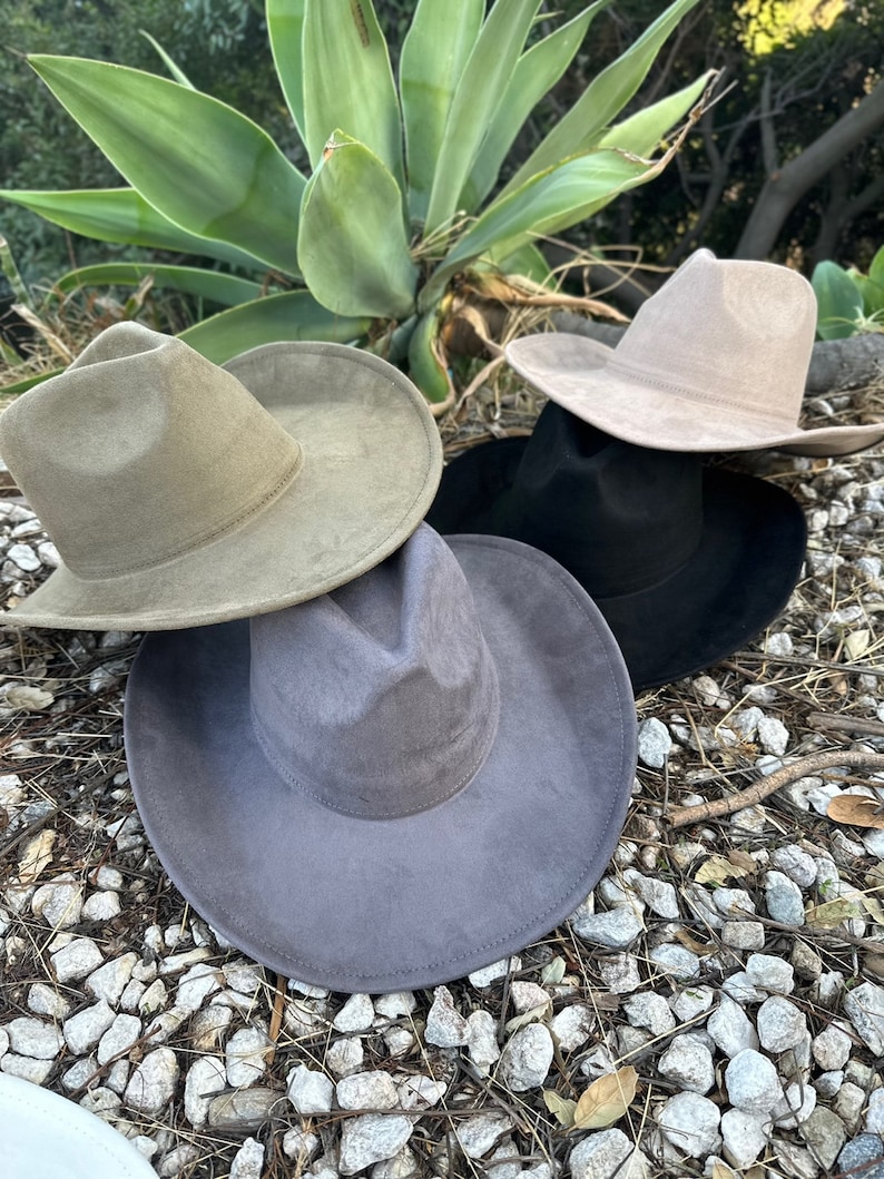 May include: A collection of four wide-brimmed fedora hats in olive green, light beige, black, and dark grey. Each hat features a structured crown with a central crease and a wide, flat brim. The hats are arranged in a cluster, showcasing their smooth, matte-textured finish and consistent design across all four colour variations.