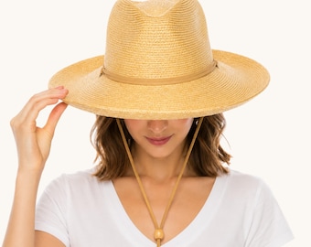 May include: A light tan straw hat with a wide brim and a brown band around the crown. The hat has a chin strap with a wooden bead. The person is wearing a white V-neck shirt.