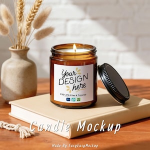 May include: A lit candle in a brown glass jar with a black lid, sitting on a beige book. The label reads "Your DESIGN here". A vase with dried plants is to the left. The words "Candle Mockup" are at the bottom.