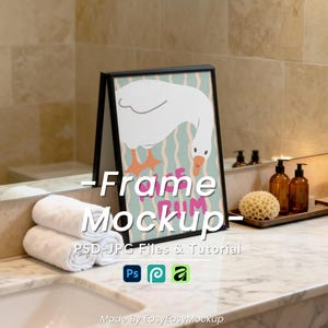May include: A framed art print featuring a white goose with orange feet and a teal background with wavy lines. The frame is black and stands on a bathroom counter next to rolled white towels and a sink.