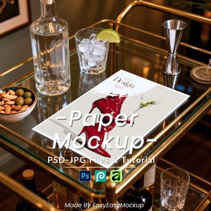 May include: A gold bar cart with a clear glass top holds a bottle of liquor, a glass of ice, and a paper mockup with a red shoe design. A bowl of nuts and olives, a cocktail shaker, and a strainer are also on the cart. The text "Paper Mockup" is visible.