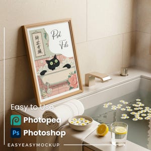 May include: A framed print featuring a black cat in a bathtub with bubbles, the text "Psd File", and Japanese characters. The print is in a light wood frame. The image is set in a bathroom with a towel, bowl of flowers, lemon, and a glass of water.