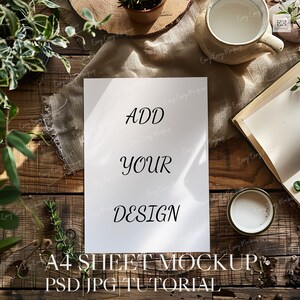 May include: A white sheet of paper with the text "ADD YOUR DESIGN" on a wooden table with plants and a cup of coffee. The text "A4 SHEET MOCKUP PSD JPG TUTORIAL" is below the paper.