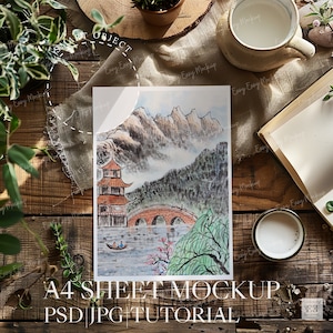 May include: A watercolor painting of a traditional Chinese landscape with mountains, a bridge, and a pagoda. The painting is on a white sheet of paper, which is on a wooden table with a book, a cup of coffee, and a plant.
