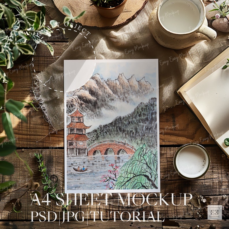 May include: A watercolor painting of a traditional Chinese landscape with mountains, a bridge, and a pagoda. The painting is on a white sheet of paper, which is on a wooden table with a book, a cup of coffee, and a plant.