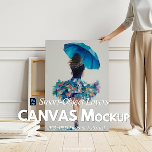 以下が含まれることがあります： 青い傘とカラフルなドレスを着た女性の絵が描かれたキャンバスモックアップ。キャンバスは人が持っています。キャンバスのテキストは「Smart-Object Layers CANVAS Mockup JPG-PSD Files & Tutorial」と書かれています。