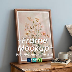 Peut inclure: Un cadre en bois clair contenant une impression d'art floral. L'œuvre présente des fleurs de style aquarelle dans des tons de rose, de jaune et de vert. Le cadre est posé sur une table de chevet en bois à côté d'une tasse, d'un livre et de lunettes. Le texte sur le cadre indique "Frame Mockup".