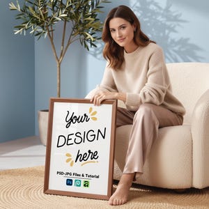 Könnte beinhalten: Ein gerahmter Druck mit dem Text "Your DESIGN here" in einem braunen Holzrahmen. Der Druck wird von einer Person gehalten, die einen beigen Pullover und eine Hose trägt und in einem cremefarbenen Stuhl sitzt. Der Hintergrund umfasst eine Pflanze und eine blaue Wand.