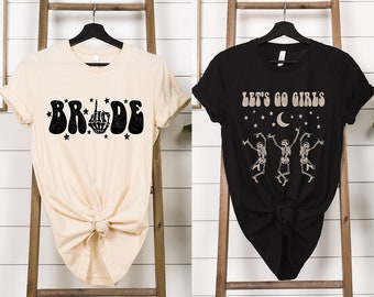Puede incluir: Dos camisetas con un diseño retro. La primera camiseta es beige con texto negro que dice "BRIDE" con un dedo medio de esqueleto. La segunda camiseta es negra con texto blanco que dice "LET'S GO GIRLS" con tres esqueletos bailando.