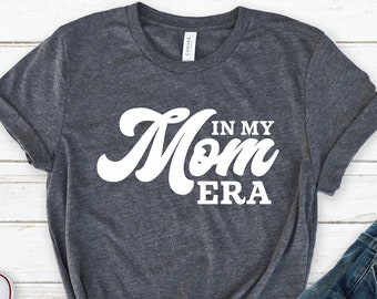 May include: A heather gray t-shirt with the text "Mom in my Era" in white lettering. The shirt is folded and laid on a white wood surface with a pair of white sneakers and a pair of blue jeans.