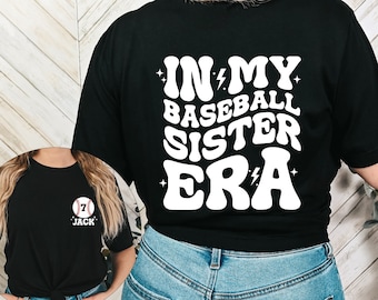 May include: Black t-shirt with white text that reads "In My Baseball Sister Era" with lightning bolts and stars. The shirt is being worn by a person with long blonde hair.