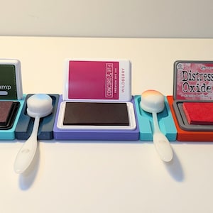 Może przedstawiać: Cztery podkładki do stempli z narzędziami do mieszania. Podkładki do stempli są w różnych kolorach i mają różne nazwy marek. Narzędzia do mieszania są z białego plastiku z gąbką na końcu.
