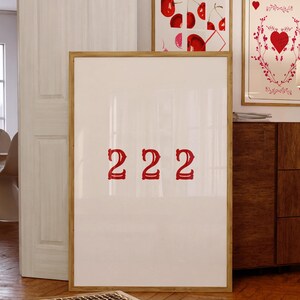 以下が含まれることがあります： 赤い文字で「2222」と書かれた白い額縁のポスター。ポスターは木製の床と木製のドレッサーのある部屋の壁にかけられている。