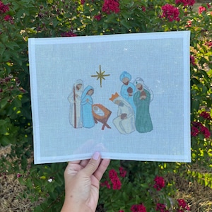May include: A rectangular white needlepoint canvas featuring a hand-painted nativity scene. The design includes figures in blue, green, and white robes surrounding a manger, with a gold star positioned above the central group on the grid.