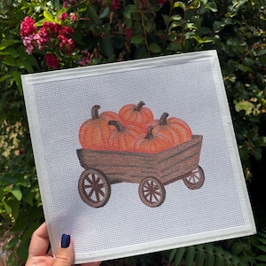 May include: A square needlepoint canvas featuring a hand-painted design of three orange pumpkins resting inside a brown wooden wagon with four spoked wheels. The design is centered on a white mesh grid with a white fabric border on all four sides.