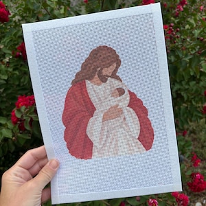 May include: A rectangular needlepoint canvas featuring a printed design of a figure with long brown hair and a beard, wearing a white robe and a red cloak, holding a small infant wrapped in white fabric against a white grid background.