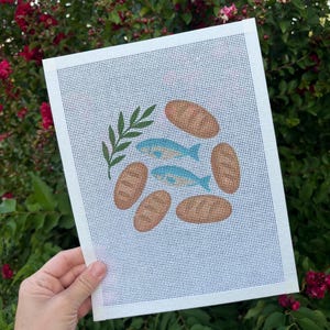 May include: A white needlepoint canvas featuring a hand-painted design of two light blue fish, five tan oval-shaped loaves of bread, and a single green leafy branch. The grid pattern of the canvas is clearly visible across the entire rectangular surface.